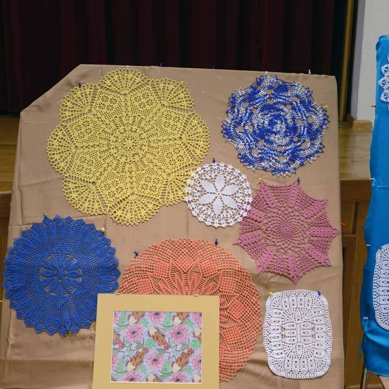 Ein brauner Tisch, bedeckt mit einer Vielzahl von gehäkelten Tischsets in verschiedenen Farben, einschließlich Gelb, Blau, Orange, Rosa und Weiß. Ein gerahmtes Bild befindet sich ebenfalls auf dem Tisch.