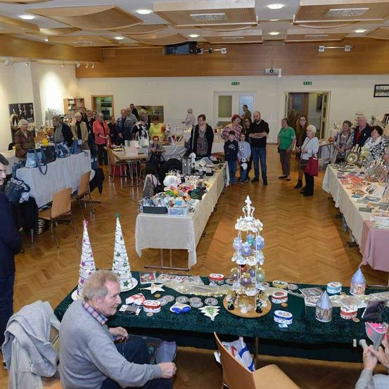 Eine Gruppe von Menschen versammelt sich in einem Raum mit Holzböden. Viele Tische mit Gegenständen sind aufgebaut, und einige Leute stöbern durch die Artikel. Einige stehen, während andere auf Stühlen sitzen. Eine Person sitzt vor einem Tisch mit Weihnachtsdekoration.