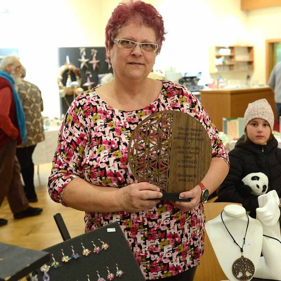 Eine Frau steht an einem Stand und hält eine Holzplakette mit einem deutschen Zitat in der Hand. Sie trägt eine Brille und ein gemustertes Oberteil. Hinter ihr sitzen eine weitere Frau und ein Kind an einem Tisch mit einem Panda-Plüschtier.