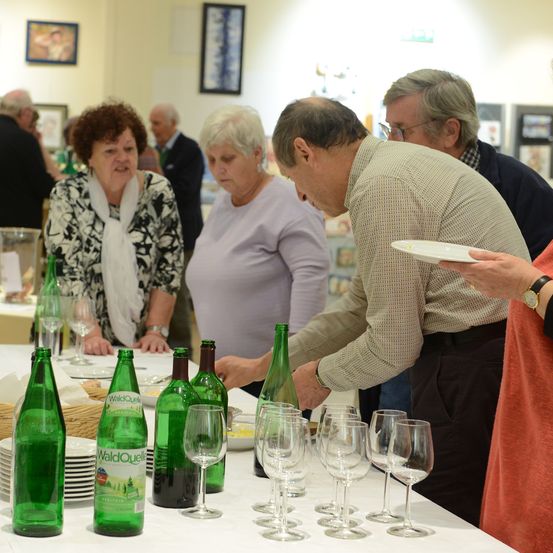 Eine Gruppe von Menschen steht um einen Tisch mit Weinflaschen und Gläsern. Ein Mann schenkt Wein in ein Glas, während andere zusehen.