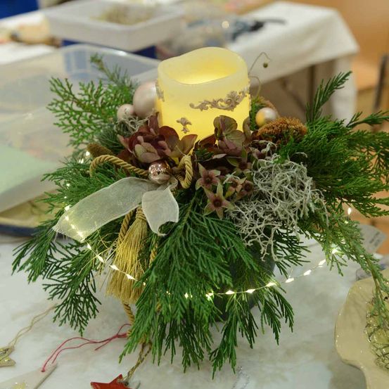 Ein Tisch-Zentralstück mit einer brennenden Kerze, umgeben von grünem Laub, Weihnachtsdekorationen und kleinen Lichtern, mit einem Band und einer Quaste.