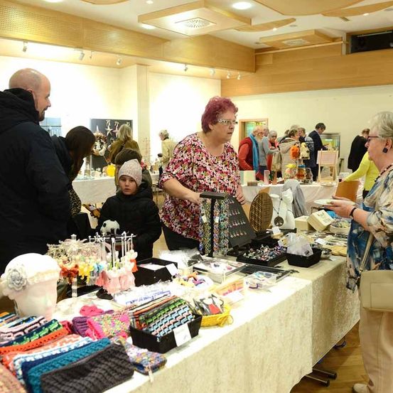 Ein Innenmarkt mit verschiedenen handgefertigten Waren im Angebot. Eine Frau mit roten Haaren betrachtet Artikel an einem Tisch. Leute stöbern durch die Produkte und unterhalten sich.
