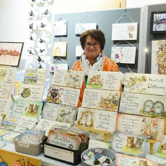 Eine Frau steht hinter einem Tisch voller verschiedener illustrierter Karten, einige mit Katzen und andere mit verschiedenen Tieren. Die Karten haben handgeschriebene Botschaften. Sie trägt eine Brille und ein Tuch und hinter ihr hängen mehrere gerahmte Kunstwerke an einer Wand.