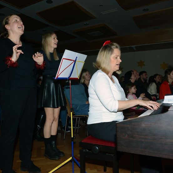Drei Frauen singen, während eine Pianistin vor einem kleinen Publikum spielt. Die Frau auf der linken Seite hat rote Dekorationen an den Handgelenken.