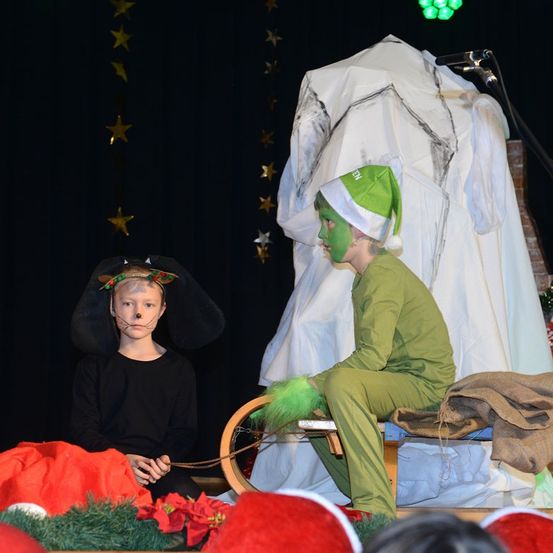Zwei Kinder sind auf der Bühne in einem Theaterstück, eines verkleidet als Maus und das andere als Elf. Ein großes weißes Kostüm steht hinter ihnen und ein rot-grüner Stoff liegt auf der Bühne.