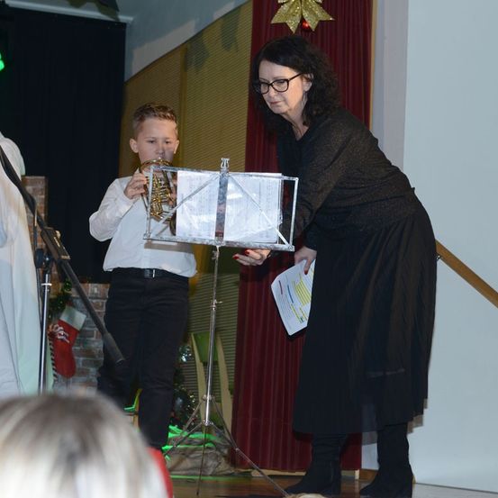 Ein junger Junge spielt Trompete auf einer Bühne, eine Frau hält einen Notenständer und Papier in der Nähe. Dahinter befindet sich ein roter Vorhang, ein Mikrofonständer und eine Ziegelwand.