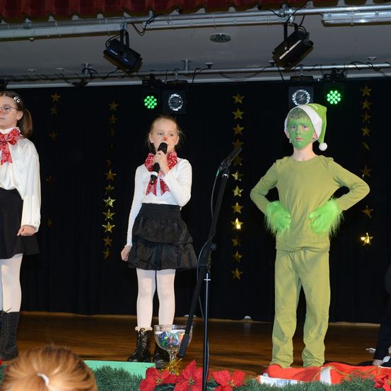 Drei Kinder auf der Bühne, eines verkleidet als Grinch, ein anderes mit einem Mikrofon und ein drittes mit einer roten Schleife. Dahinter ein Hintergrund mit Sternen und Scheinwerfern.