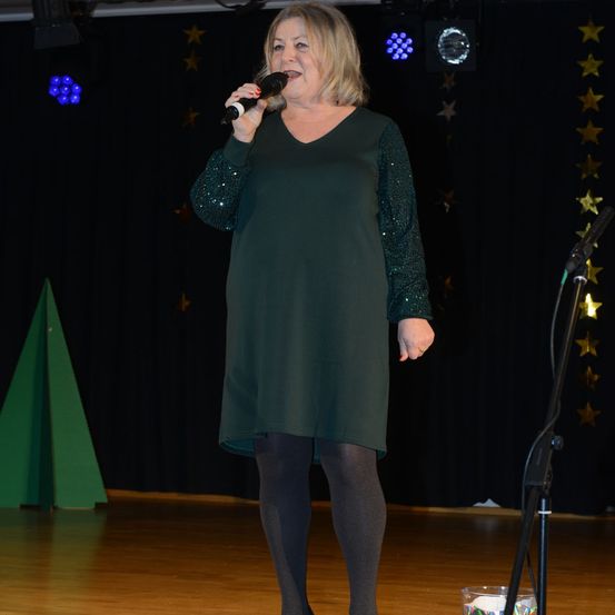 Eine Frau in einem grünen Kleid und Strumpfhosen singt auf der Bühne mit einem Mikrofon in der Hand. Die Bühne hat einen Holzboden und einen Hintergrund mit Sternen und einem grünen Baum.