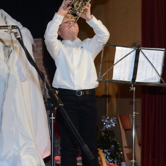 Ein junger Junge in einem weißen Hemd und schwarzen Hosen spielt Trompete auf einer Bühne. Ein Notenständer steht vor ihm.