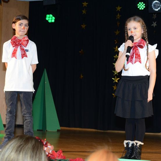 Zwei Kinder in festlicher Kleidung stehen auf der Bühne, eines hält ein Mikrofon. Der Junge hat einen roten Schal um den Hals und das Mädchen hat geflochtene Haare. Sie führen wahrscheinlich ein Weihnachtsspiel auf.