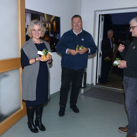 Drei Menschen stehen in einem Raum mit gefliesten Böden, ein Mann hält kleine Pflanzen und eine Frau hält eine Pflanze mit einem Schild. Ein Poster mit den Worten D&D Cocktailbar ist an der Wand. Ein anderer Mann steht in der Nähe der offenen Tür.