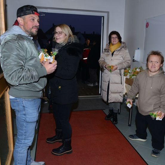 Vier Personen stehen in einem Raum mit einem roten Teppich. Ein Mann und eine Frau halten einen Snack, während die anderen beiden sie ansehen. Der Raum hat eine weiße Wand und eine Tür, die nach draußen führt.