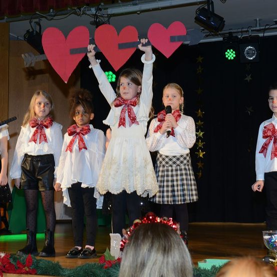 Mehrere Kinder auf der Bühne tragen Schleifen und halten einen Stock mit Herzen, ein Kind hält ein Mikrofon und ein anderes hebt die Hände.