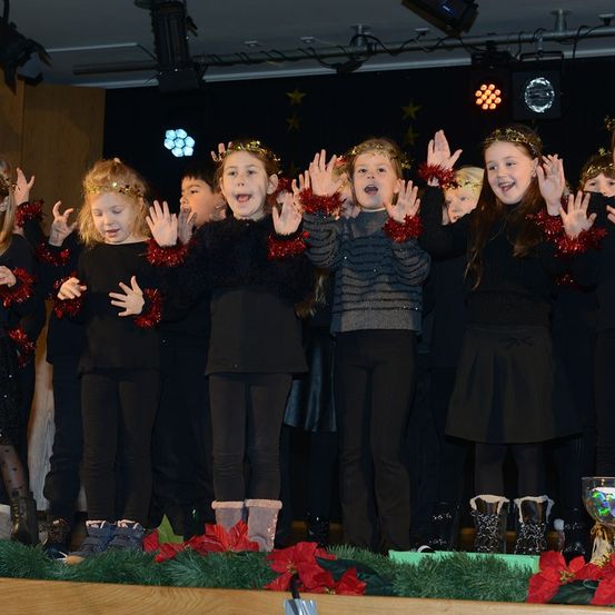 Eine Gruppe junger Kinder in schwarzen Kleidern mit goldenen Stirnbändern und rotem Tinsel steht auf der Bühne, lächelt und winkt. Sie scheinen ein Weihnachtslied vorzutragen.