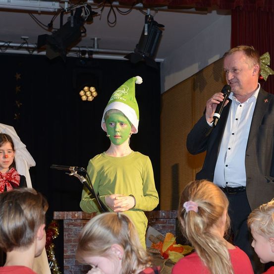Ein Junge, der als Elf verkleidet ist, spricht in ein Mikrofon, während ein Mann neben ihm spricht. Einige Kinder sitzen in der vorderen Reihe und beobachten den Mann. Scheinwerfer und Kabel sind an der Decke sichtbar.