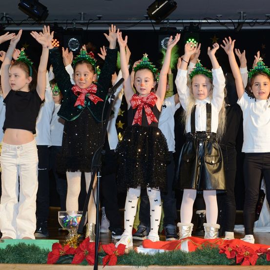 Eine Gruppe von Kindern, gekleidet in festlicher Kleidung, tritt auf der Bühne auf, mit roten Weihnachtssternen und Weihnachtsbäumen auf dem Boden. Einige tragen schwarze Kleider und weiße Stiefel, andere weiße Oberteile und schwarze Hosen.