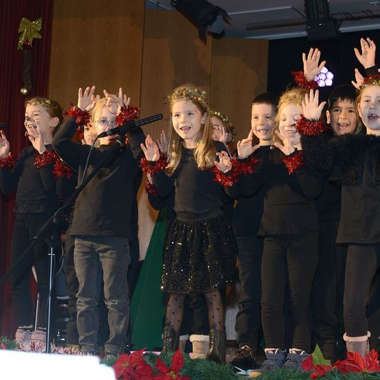 Eine Gruppe von Kindern in schwarzen Kleidern steht auf der Bühne und singt mit Mikrofonen. Sie tragen Weihnachtsdekorationen und scheinen fröhlich zu sein.