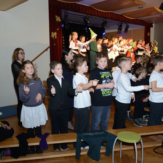 Eine Gruppe von Kindern und Erwachsenen in einem Raum, einige mit Hüten, stehen und sitzen auf Bänken. Eine Person mit einem grünen Hut steht auf der Bühne mit einem Mikrofon, und andere treten auf.