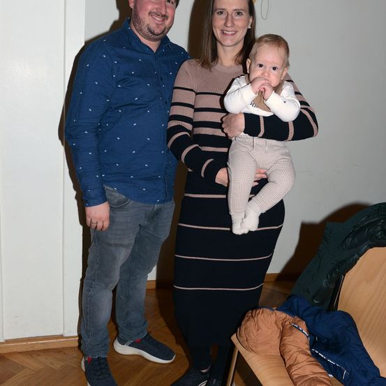 Ein Mann und eine Frau mit einem Baby in einem Wohnzimmer, lächelnd und posierend für ein Foto.