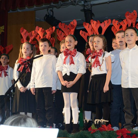 Eine Gruppe von Kindern mit roten Schleifen und Rentierkopfbedeckungen singt auf der Bühne, mit einem Mikrofon und Weihnachtsdekorationen.