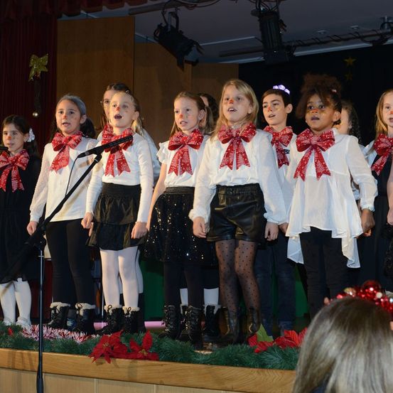 Eine Gruppe junger Mädchen mit roten Schleifen und weißen Blusen singt auf einer Bühne, die mit grünen Pflanzen und roten Blumen geschmückt ist.