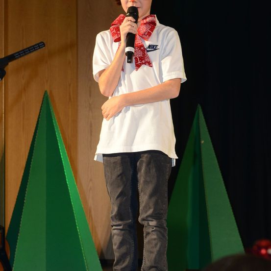 Ein Junge in einem weißen T-Shirt mit einem roten Schal um den Hals steht auf der Bühne und hält ein Mikrofon. Hinter ihm ist ein hölzerner Hintergrund und zwei grüne dreieckige Requisiten.