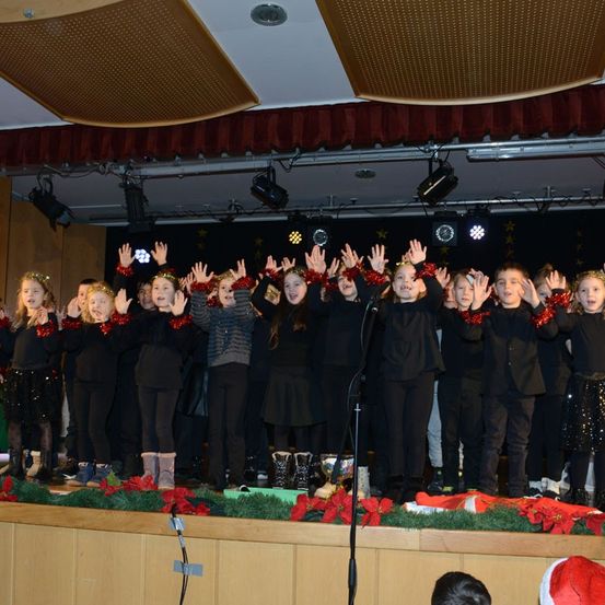 Ein Chor junger Kinder in schwarzer Kleidung tritt auf einer Bühne mit Weihnachtsdekorationen und einem Weihnachtsmannhut auf, über der Bühne hängen Scheinwerfer.