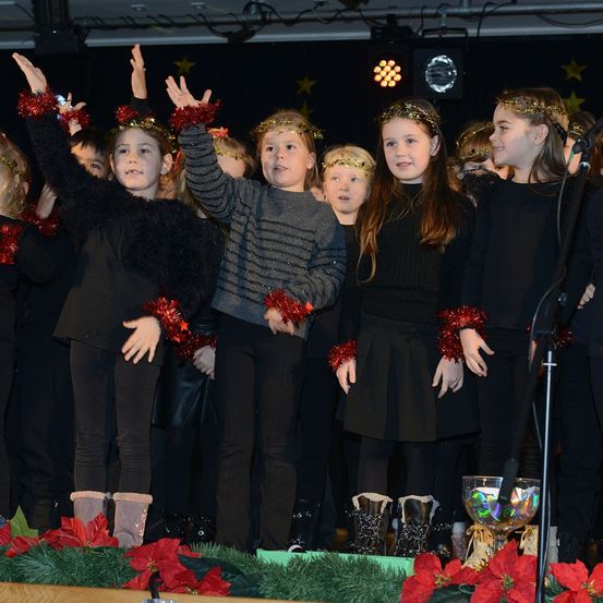 Eine Gruppe von Kindern, die in schwarze und rote Ornamente gekleidet sind, tritt auf der Bühne auf, lächelt und hebt die Hände. Die Bühne ist mit roten Weihnachtssternen und grüner Girlande geschmückt. Ein Mikrofon steht vor den Kindern.