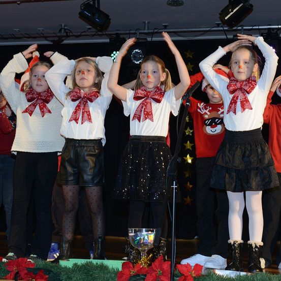 Mehrere Kinder in übereinstimmenden weißen Hemden mit roten Schleifen und schwarzen Röcken treten auf der Bühne mit Mikrofonen auf, beleuchtet von Bühnenlichtern und umgeben von roten Christsternen.
