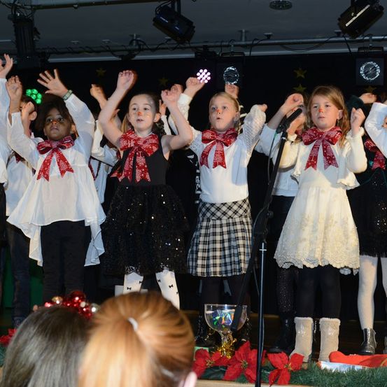 Eine Gruppe von Kindern in festlicher Kleidung tritt auf der Bühne auf und hebt die Hände. Die Bühne ist mit Weihnachtsdekorationen geschmückt, und Scheinwerfer beleuchten die Darsteller.