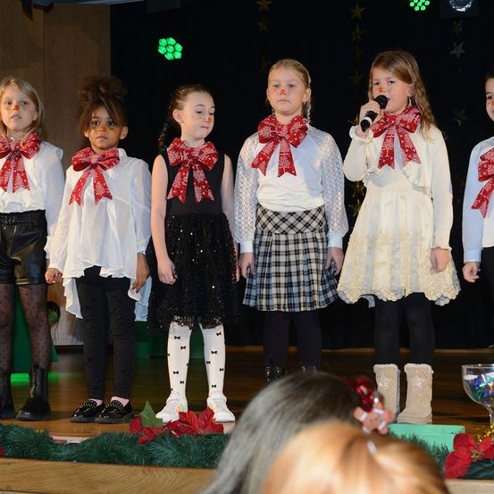 Eine Gruppe junger Mädchen mit roten Schleifen steht auf der Bühne und singt. Eine von ihnen hält ein Mikrofon. Sie tragen weihnachtliche Outfits. Dahinter befindet sich eine Bühne mit grünen Blättern und roten Blumen.