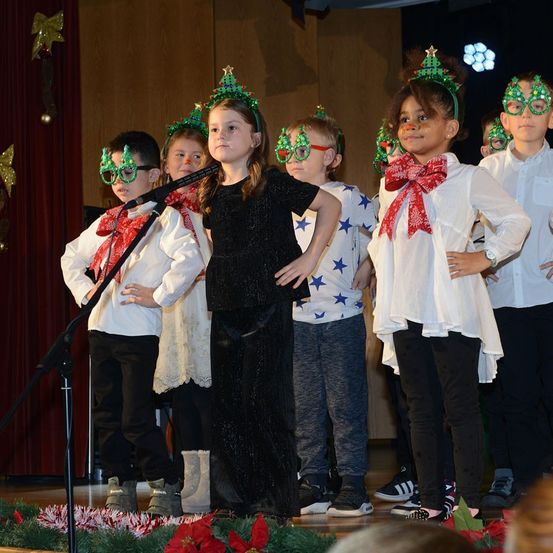 Eine Gruppe von Kindern auf der Bühne, gekleidet in festlichem Outfit, singen ein Weihnachtslied. Eines der Mädchen hält ein Mikrofon. Sie tragen Weihnachtskopfbänder und weiße Schleifen.