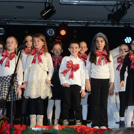 Eine Gruppe von Kindern mit roten Schleifen um den Hals steht mit Mikrofonen auf der Bühne und singt wahrscheinlich bei einer Schulveranstaltung.