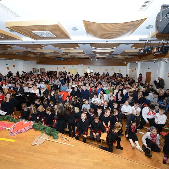 Eine große Menschenmenge versammelt sich in einem Raum für eine festliche Veranstaltung, mit Kindern in schwarzen Kostümen und Kränzen, Erwachsenen auf hölzernen Böden sitzend und einer dekorierten Bühne mit Klavier und Grünpflanzen. Die Decke ist mit Sternenmustern und Beleuchtungskörpern versehen.