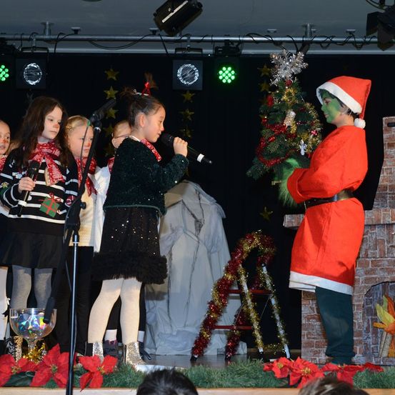 Eine Gruppe junger Mädchen auf der Bühne singt während einer Weihnachtsaufführung. Ein Junge, der als Weihnachtsmann gekleidet ist, steht neben einem Weihnachtsbaum.