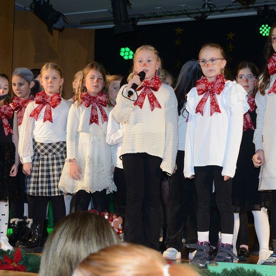 Eine Gruppe von Kindern mit roten Schals und weißen Outfits steht auf der Bühne und singt mit einem Mikrofon. Sie haben rote Nasen und scheinen zu singen. Die Bühne ist mit Weihnachtslichtern dekoriert und es gibt Zuschauer im Publikum.