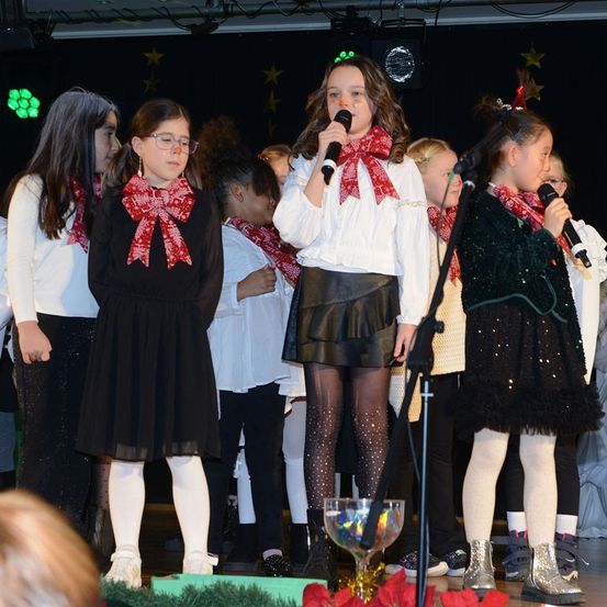 Eine Gruppe junger Mädchen in festlicher Kleidung steht auf der Bühne und hält Mikrofone. Ein Mädchen in einem weißen Hemd mit rotem Schleifenband singt, während andere zuhören. Sie sind mit Schals und Stiefeln geschmückt, und die Bühne ist mit roten Weihnachtssternen und einem großen Glasornament dekoriert.