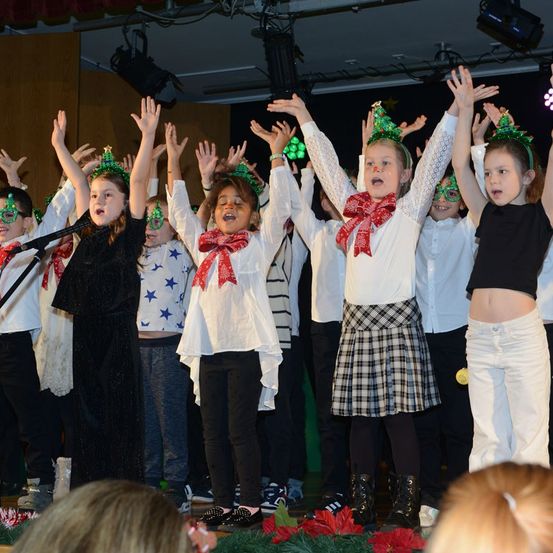 Eine Gruppe von Kindern tritt auf der Bühne auf, trägt Weihnachtskopfbänder und hält die Hände hoch. Einige tragen weiße Kleider mit schwarzen Röcken und Stiefeln. Ein Mikrofon steht vor ihnen, und der Hintergrund ist mit Weihnachtsbäumen und Lichtern geschmückt.