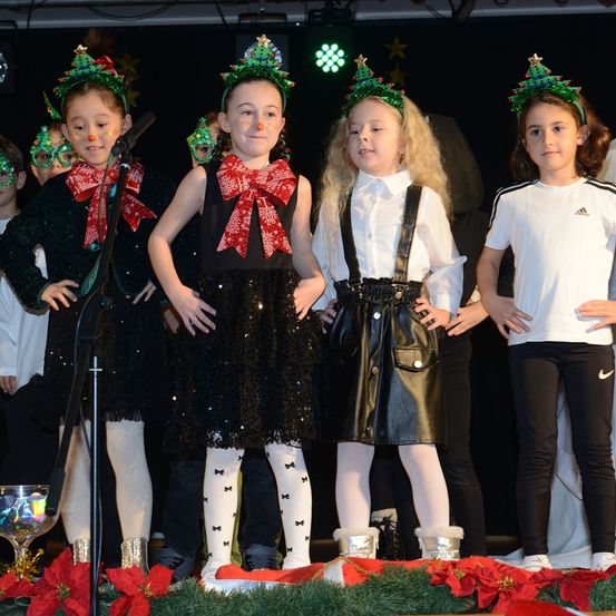 Eine Gruppe junger Mädchen in weihnachtlich geschmückten Kostümen steht auf der Bühne. Sie tragen Kopfbänder mit Weihnachtsbäumen und roten Schleifen. Ein Mikrofon steht vor ihnen. Auf der Bühne sind rote Blumen.