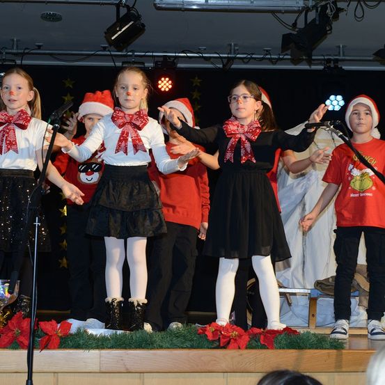 Eine Gruppe von Kindern auf der Bühne, gekleidet in festlicher Kleidung, führt ein weihnachtliches Theaterstück mit einem festlichen Hintergrund auf, der mit Weihnachtssternen geschmückt ist.