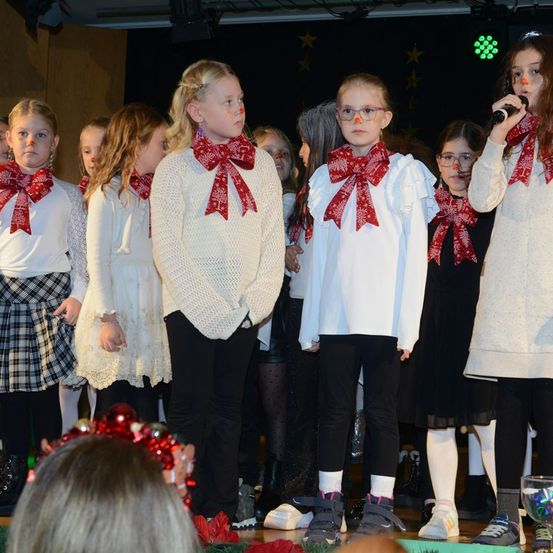 Eine Gruppe junger Mädchen mit roten Schleifen an ihren Kleidern steht auf der Bühne, wahrscheinlich bei einer Schulveranstaltung. Sie tragen weiße Kleider und schwarze Hosen, einige tragen Brillen. Ein Mädchen in der Mitte hält ein Mikrofon und scheint zu singen.