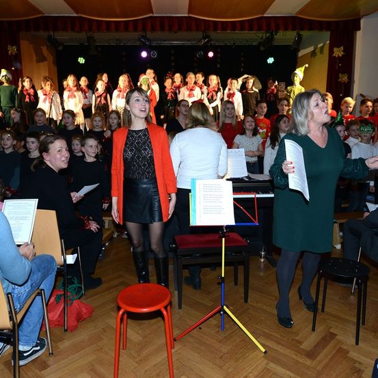 Eine Choraufführung in Innenräumen mit einer Gruppe von Kindern und Erwachsenen, die singen, wobei zwei Frauen vor ihnen Notenblätter halten. Dahinter spielt eine Pianistin an einem Klavier. Das Publikum sitzt auf Stühlen, einige tragen festliche Mützen.
