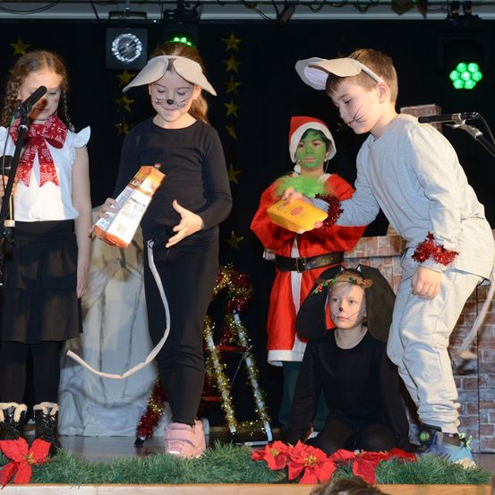 Eine Gruppe von Kindern tritt auf der Bühne mit Weihnachtsdekorationen auf. Ein Kind, verkleidet als Grinch, hält eine gelbe Box. Ein weiteres Kind in einem Weihnachtsmann-Kostüm steht hinter ihnen. Ein Mädchen in einer Katzen-Verkleidung kauert in der Nähe.