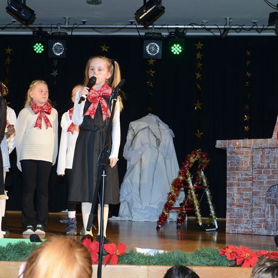 Ein junges Mädchen steht auf der Bühne und singt in ein Mikrofon mit einer Schleife um den Hals. Hinter ihr stehen andere Kinder mit Schleifen, und ein weißes Gewand ist über eine Leiter gelegt. Die Bühne ist mit Weihnachtssternen und einem Backsteinkamin dekoriert. Scheinwerfer sind an der Decke montiert.