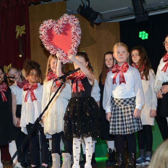 Eine Gruppe von Kindern auf der Bühne, eines hält ein großes Herz und trägt Schleifen und weiße Schals. Sie treten für ein Publikum auf.