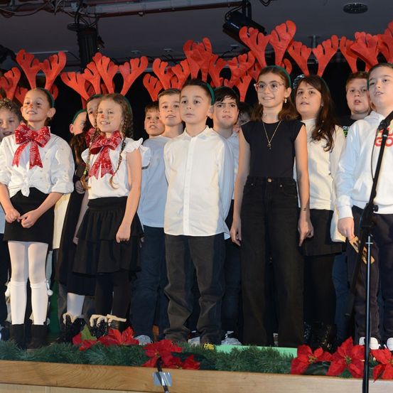 Eine Gruppe von Kindern steht auf der Bühne mit roten Rentierkopfbändern und roten Schleifen. Sie singen mit einem Mikrofon vor sich.