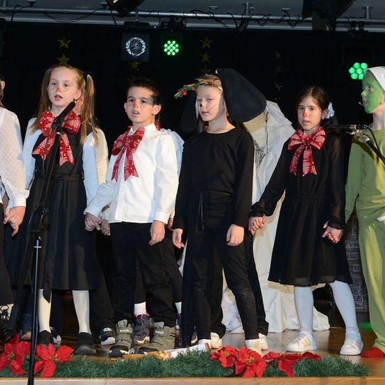 Eine Gruppe von Kindern in Kostümen tritt mit Mikrofonen auf der Bühne auf, die mit roten Blumen geschmückt ist. Sie haben rote Schleifen an ihren Kleidern.