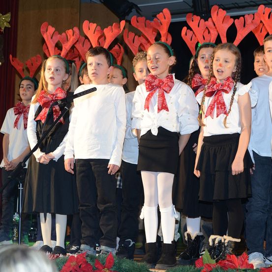 Ein Chor von Kindern in Weihnachtskleidung, die rote Stirnbänder mit Rentierhörnern tragen, steht auf einer Bühne und singt. Sie haben rote Bögen um den Hals.