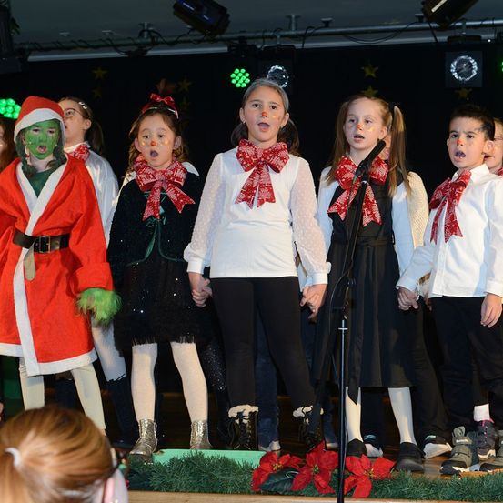Eine Gruppe von Kindern in Weihnachtskostümen tritt auf der Bühne auf, einer als Grinch verkleidet. Sie singen in ein Mikrofon, mit Christsternen und grünen Pflanzen im Vordergrund.