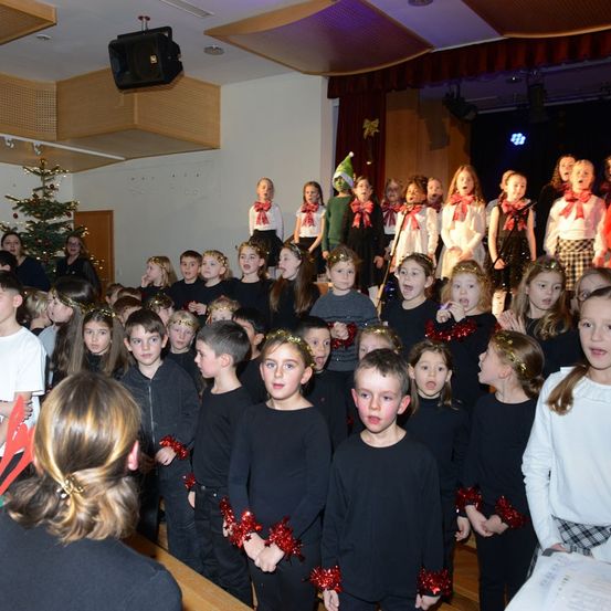 Ein Chor von Kindern in schwarzen Outfits und roten Stirnbändern singt auf der Bühne, mit einem Weihnachtsbaum im Hintergrund. Einige Kinder haben rote Schleifen um den Hals.